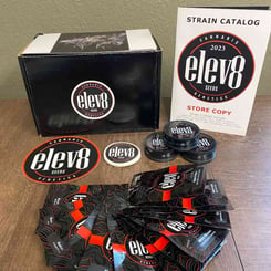 A collection of Elev8 Seeds promotional materials, including seed packages, stickers, a plastic container, and a 2023 strain catalog featuring the highly acclaimed elev8 Bakers Dozen, are displayed on a wooden surface.