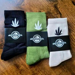 Three NASC Socks in black, green, and beige, each featuring a white cannabis leaf design and a label reading "North Atlantic Seed Co.," are displayed on a wooden surface by NASC.