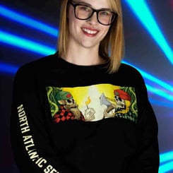 A person with shoulder-length hair and glasses smiles at the camera. They are wearing a black NASC Jack & Jill Long Sleeve Shirt [LIMITED], decorated with a graphic featuring two skeletons. Blue laser lights are in the background.