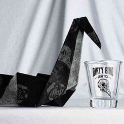 A black origami bird is placed next to a shot glass with the text "DIRTY BIRD GENETICS" on it, set against a white draped background, showcasing the Dirty Bird Outdoor Box Set - Half Packs (F).