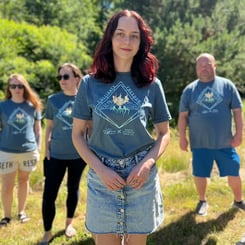 Four people standing outdoors on grass, one woman in the foreground wearing a denim skirt and a Cultivate Freedom T-Shirt, with three others in similar outfits in the background. Trees and sunlight fill the backdrop as they celebrate Pride Month, sparking the rainbow with their joyful presence.