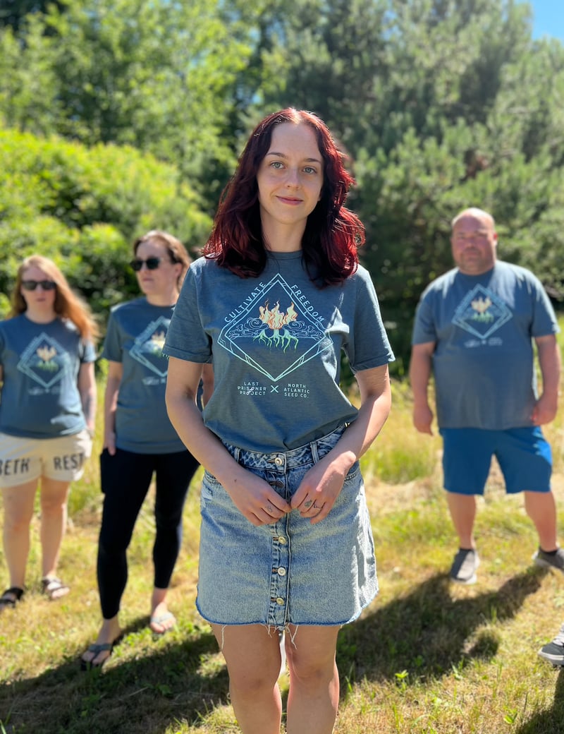 Four people standing outdoors on grass, one woman in the foreground wearing a denim skirt and a Cultivate Freedom T-Shirt, with three others in similar outfits in the background. Trees and sunlight fill the backdrop as they celebrate Pride Month, sparking the rainbow with their joyful presence.