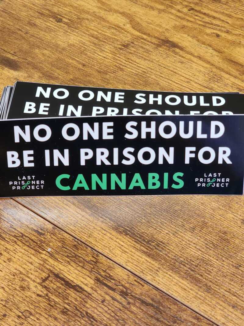 Stack of Last Prisoner Project Stickers with the text "NO ONE SHOULD BE IN PRISON FOR CANNABIS" and the Last Prisoner Project logo on a wooden surface.